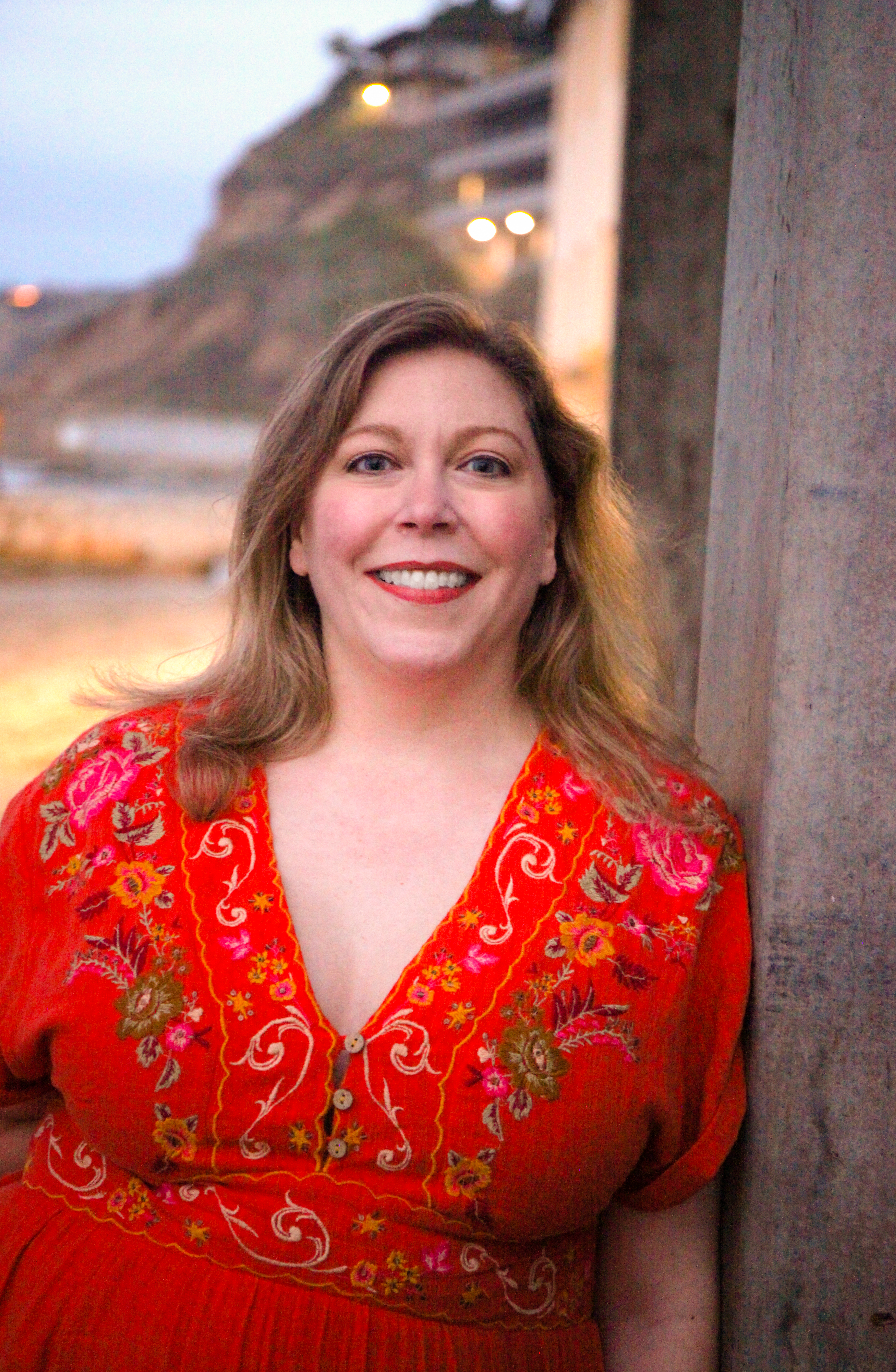 Smiling woman in an orange dress leaning against a pier at dusk.